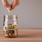 Hand placing a coin into a jar labeled Budget, representing long-term parking lot maintenance budgeting